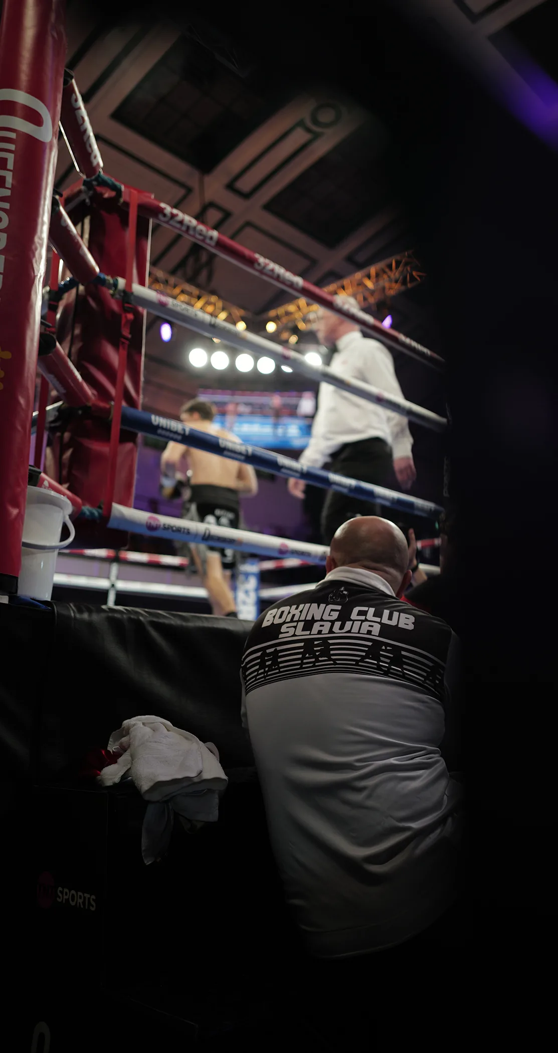 A cornerman watching from ringside as two boxers work under the arena lights
