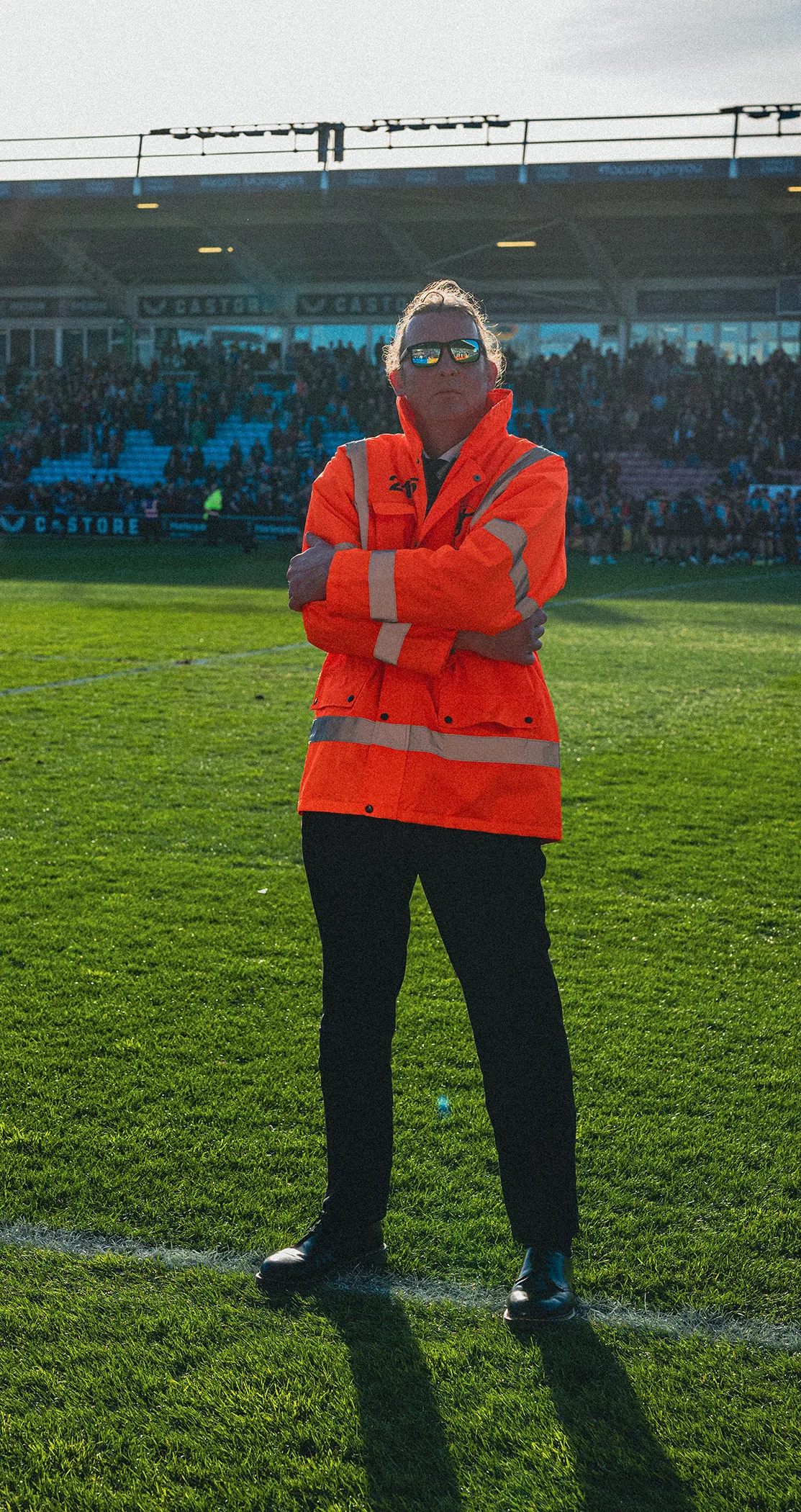 A steward in an orange hi-vis jacket standing arms folded at the pitch edge in low sun