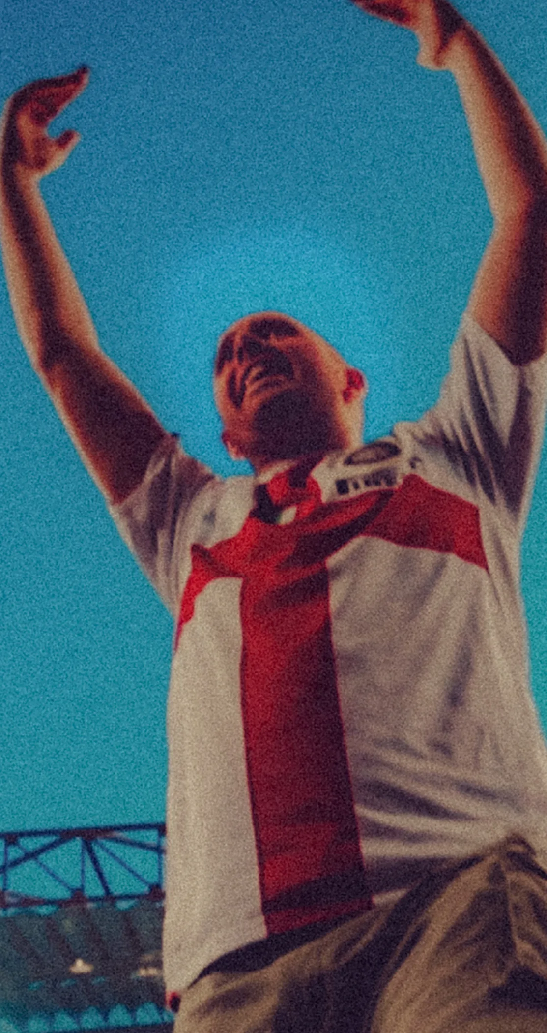 A fan in a white shirt with a red cross, arms raised, seen from below against the stadium roof, San Siro